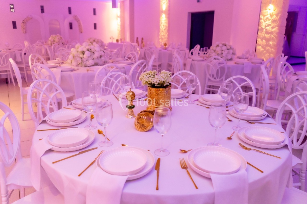 Salle de réception décorée avec des tables rondes dressées, nappes blanches, couverts dorés et arrangements floraux.