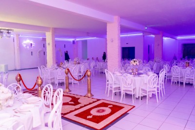 Salle de réception décorée avec des tables rondes, nappes blanches, chaises élégantes et éclairage violet tamisé.