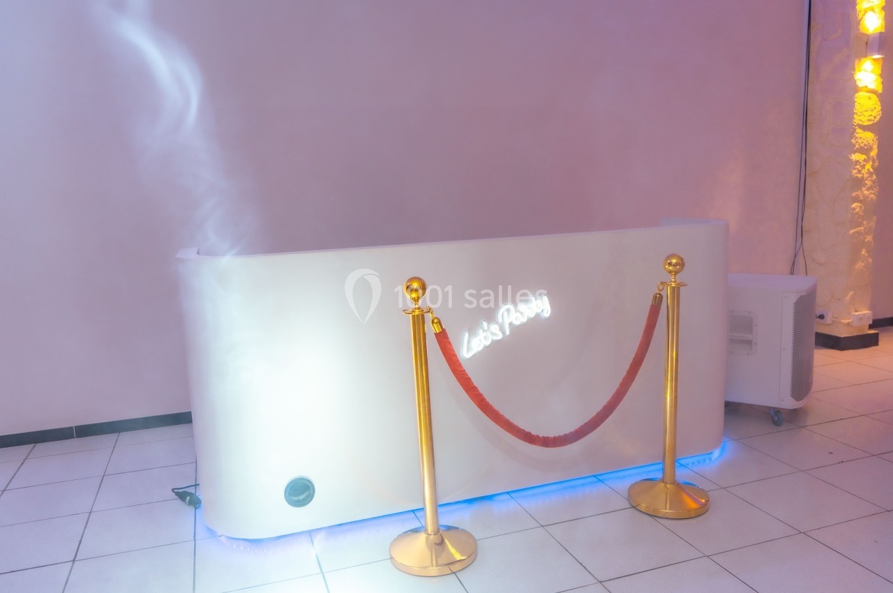 Comptoir blanc lumineux avec inscription ’Let's Party’, entouré de poteaux dorés et d'une corde rouge, dans une pièce…