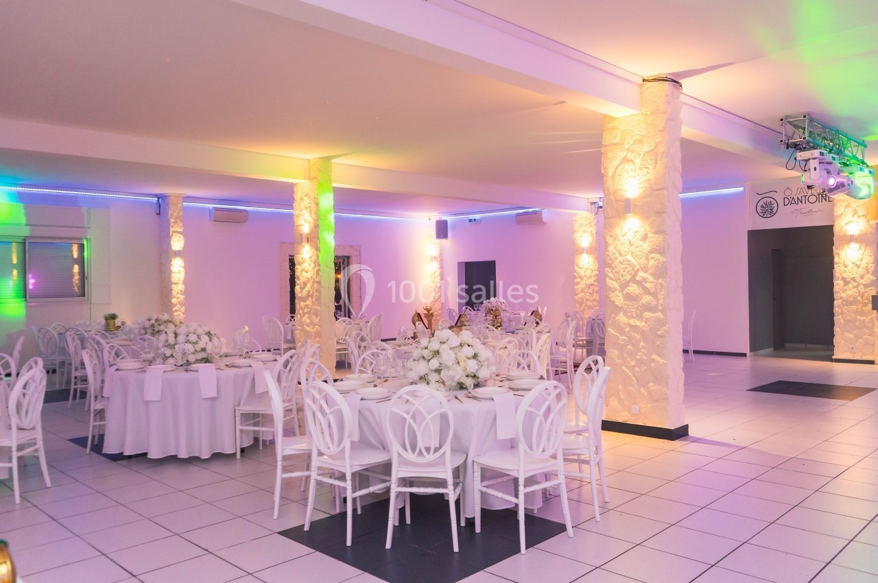 Salle de réception lumineuse avec tables rondes dressées, chaises blanches et décorations florales blanches.