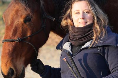 Femme souriante tenant les rênes d'un cheval brun dans un environnement extérieur boisé.