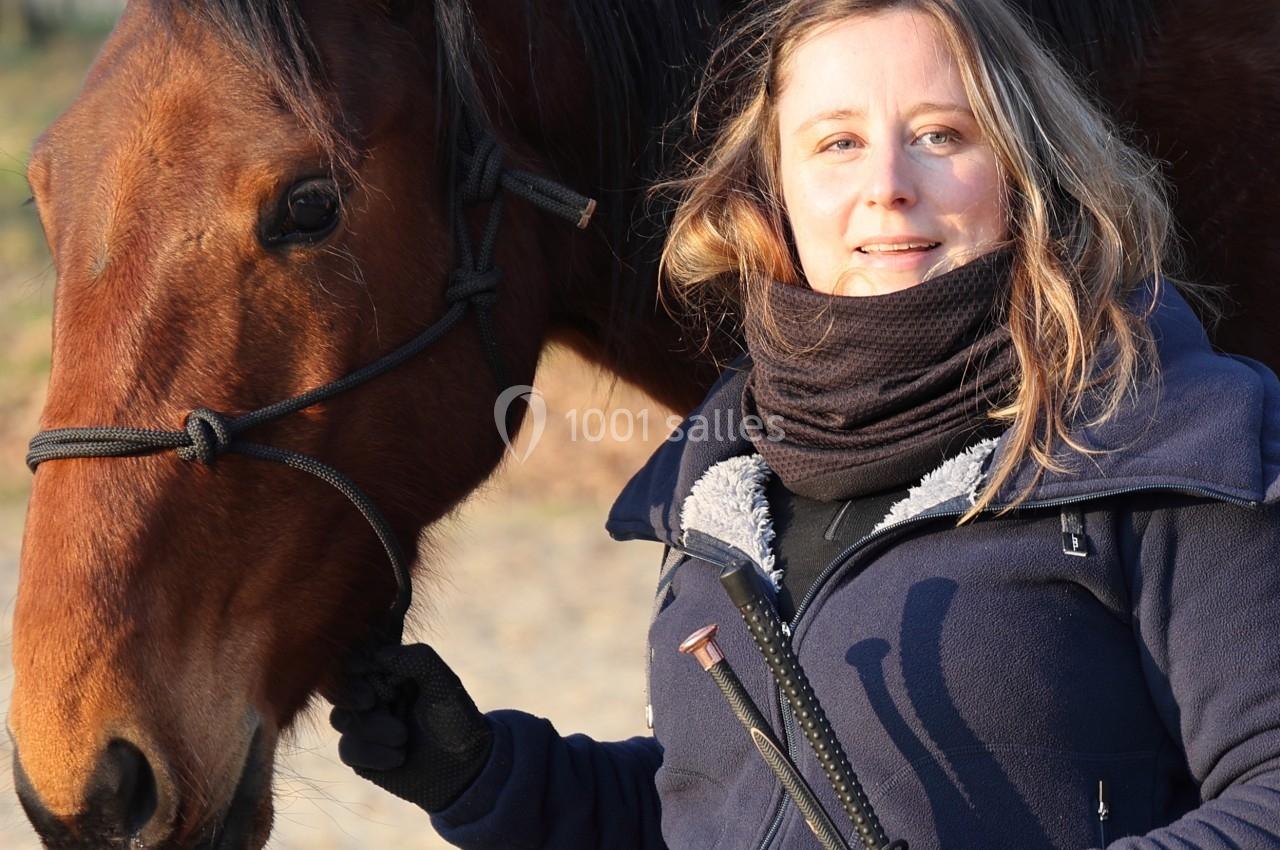 Femme souriante tenant les rênes d'un cheval brun dans un environnement extérieur boisé.