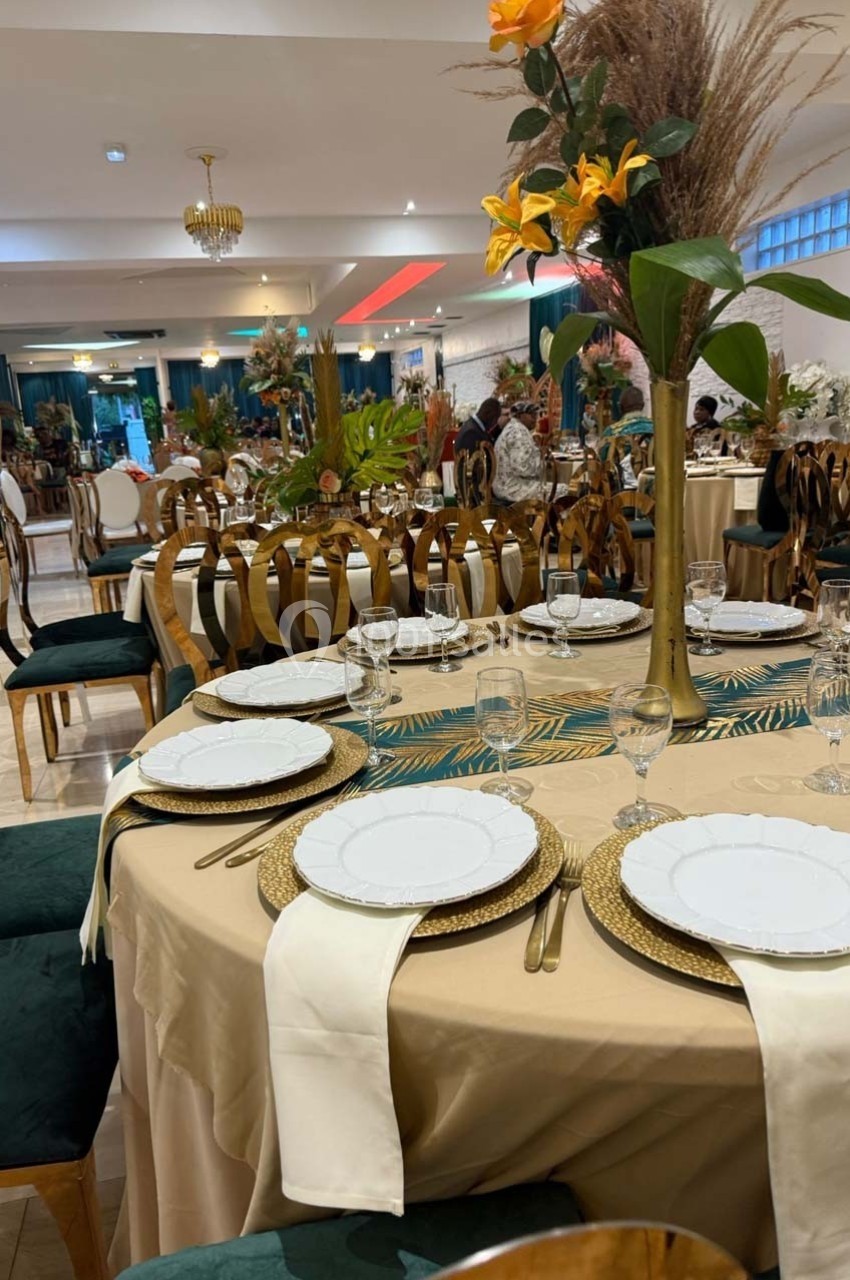 Salle de réception décorée avec des tables rondes élégantes, nappes beiges, assiettes blanches et centres de table fleuris.
