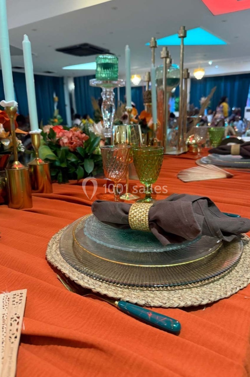 Table élégamment dressée avec nappe orange, assiettes superposées, serviette grise et verres colorés dans un décor festif.