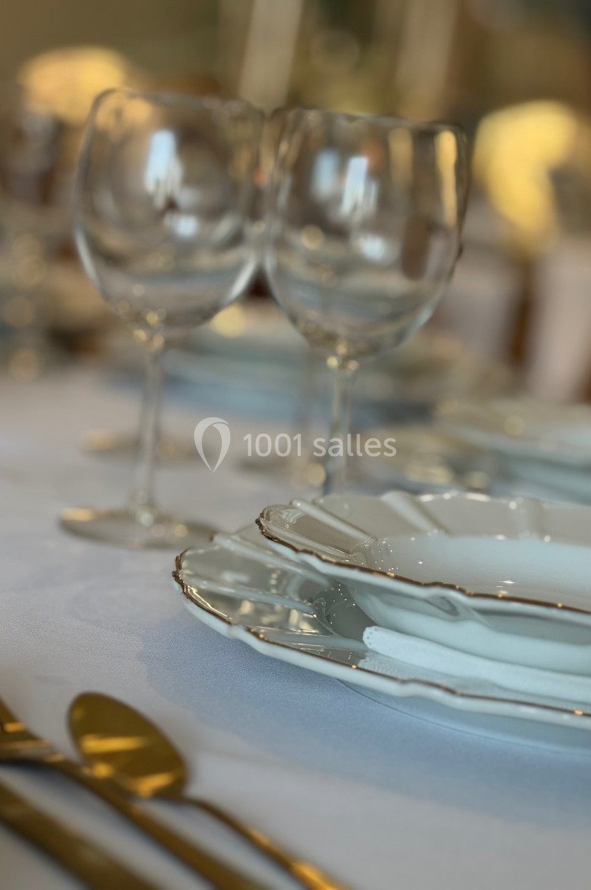 Table dressée avec des assiettes blanches bordées d'or, verres à pied et couverts dorés sur une nappe blanche.
