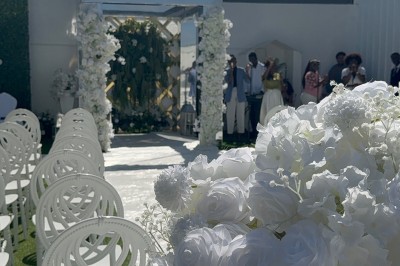 Allée centrale décorée de fleurs blanches menant à une arche florale, entourée de chaises blanches en extérieur.
