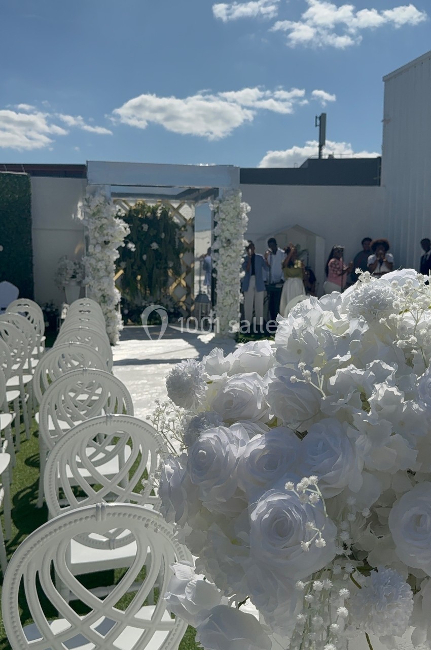 Allée décorée de chaises blanches et de fleurs blanches menant à une arche florale lors d'une cérémonie en extérieur.