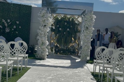 Allée centrale décorée de fleurs blanches menant à une arche florale, entourée de chaises blanches en extérieur.