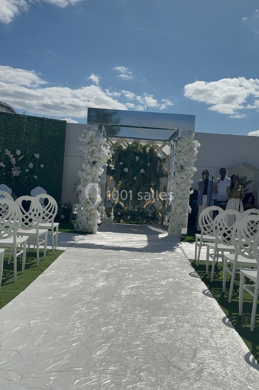 Allée centrale décorée de fleurs blanches menant à une arche florale, entourée de chaises blanches en extérieur.