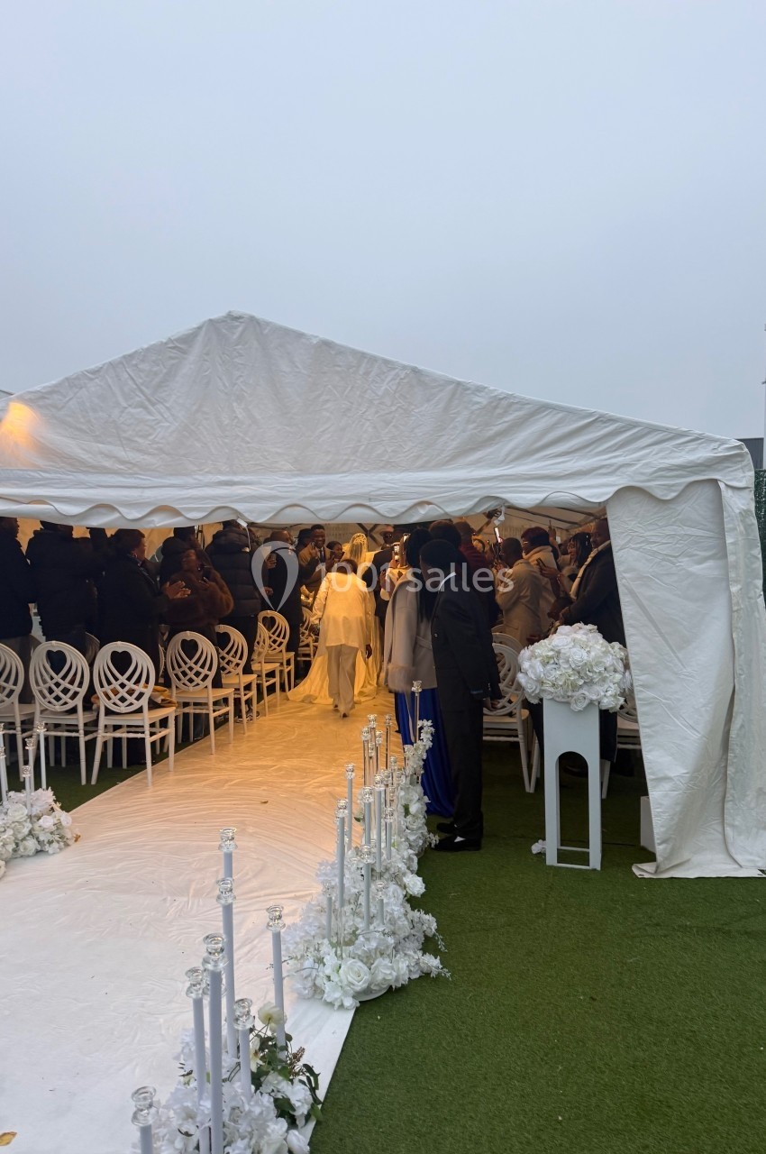 Cérémonie en plein air sous une tente blanche, avec des invités assis et un tapis blanc décoré de fleurs.
