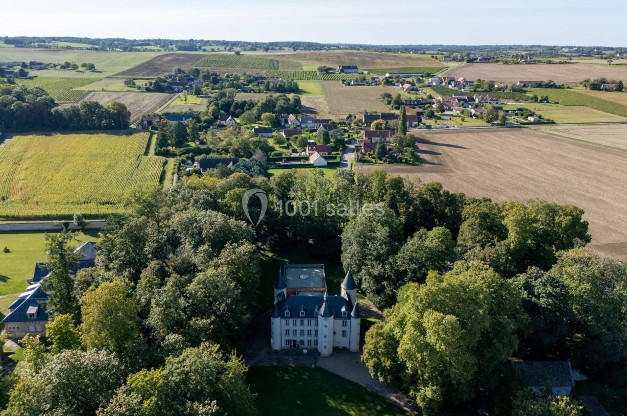 Vue aérienne d'un château entouré d'arbres, avec des champs et un village en arrière-plan.
