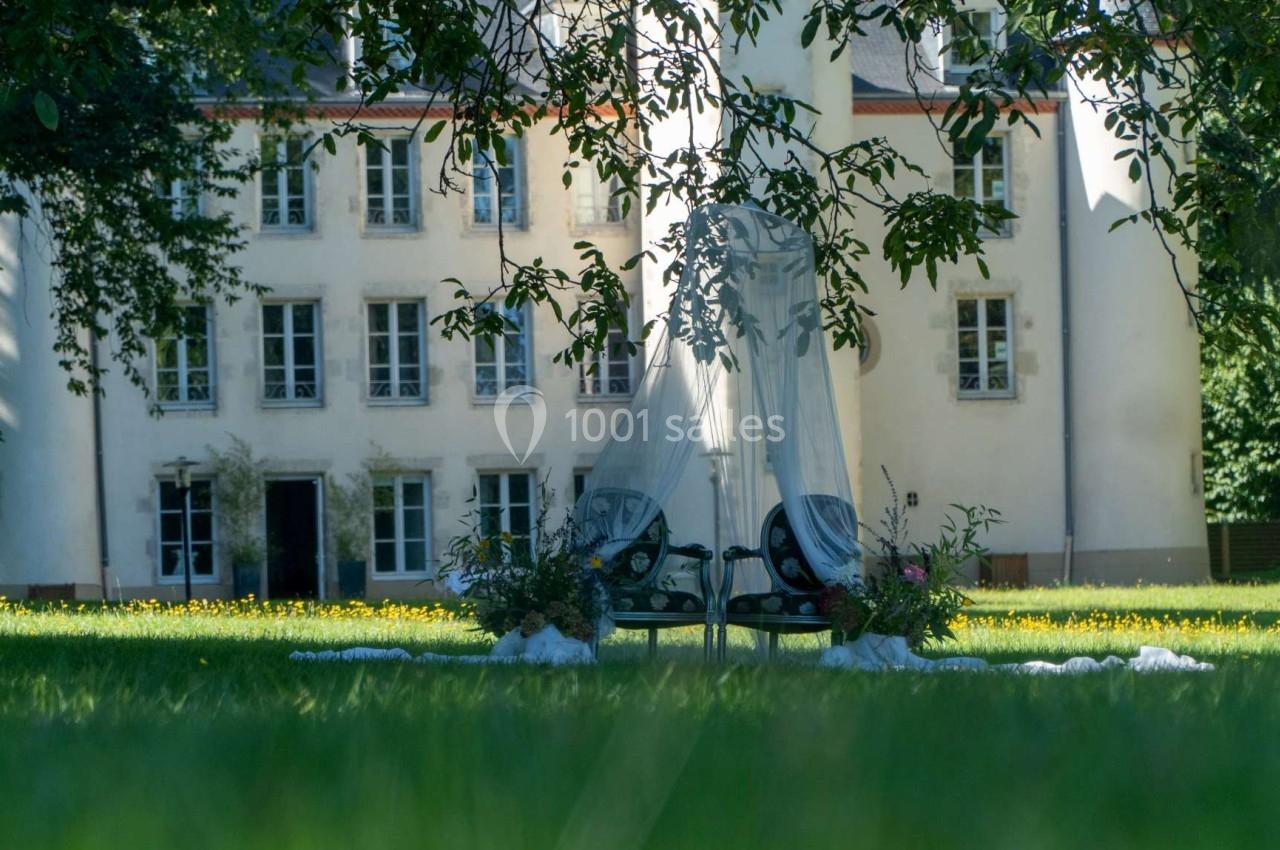Deux chaises longues avec un voile blanc, entourées de fleurs, installées sur une pelouse devant un bâtiment ancien.