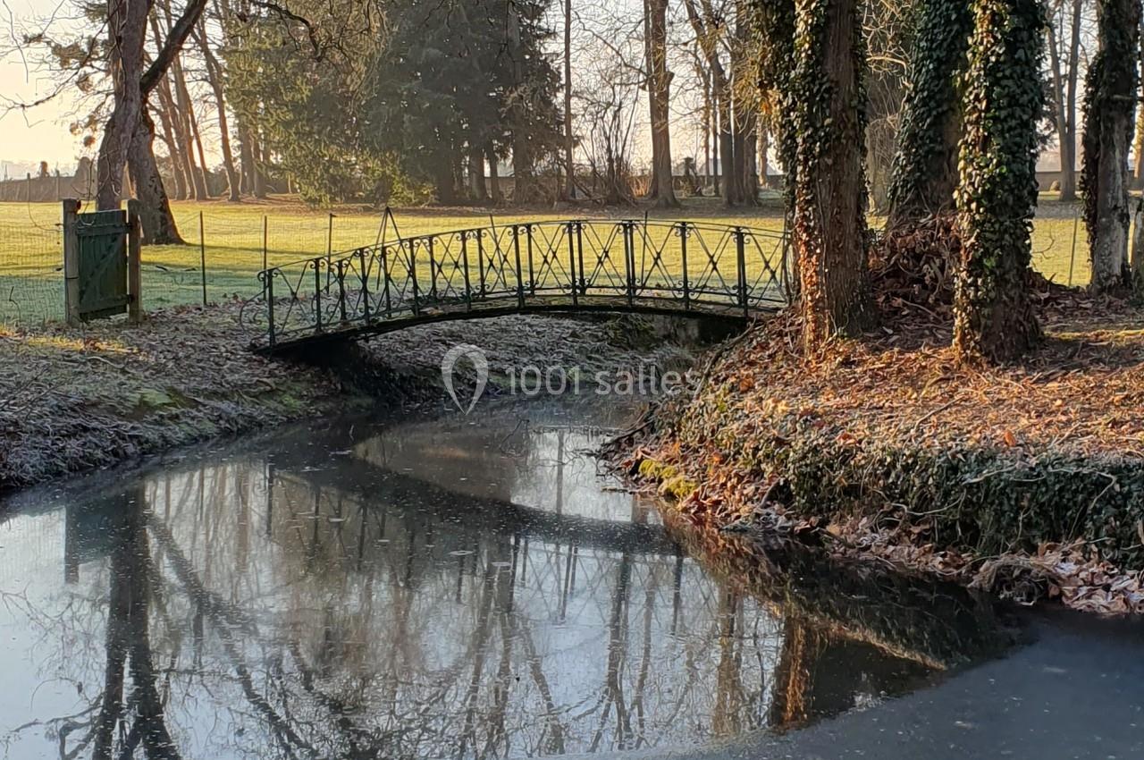 Petit pont en métal traversant un ruisseau entouré d'arbres en hiver, avec un champ en arrière-plan.