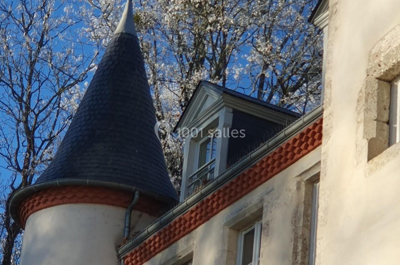 Toiture d'un bâtiment ancien avec une tour conique et des fenêtres, entourée d'arbres en fleurs sous un ciel bleu.