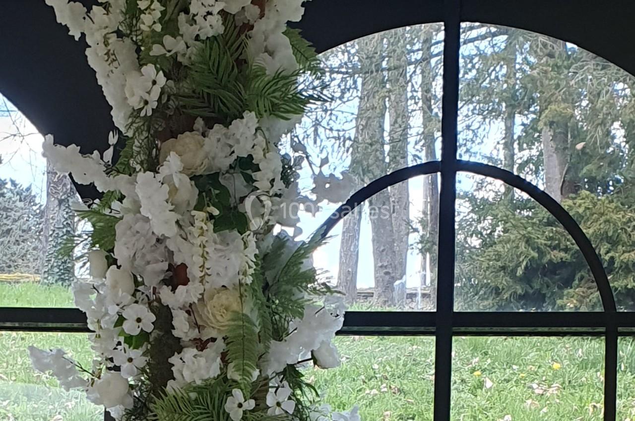 Arrangement floral blanc et vert devant une fenêtre cintrée donnant sur un jardin avec des arbres.