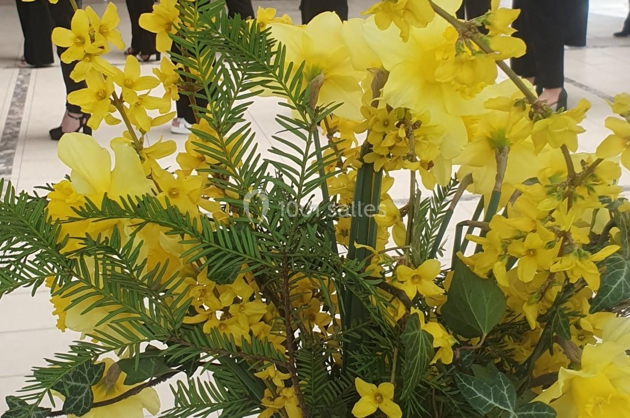Un bouquet de fleurs jaunes dans un vase en verre, avec des personnes debout en arrière-plan dans une salle lumineuse.