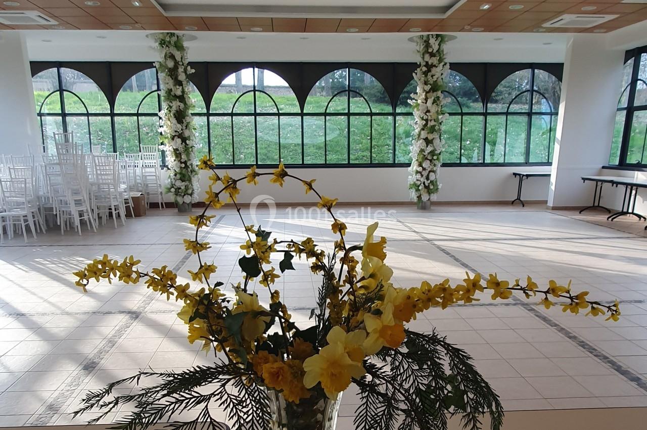 Salle lumineuse avec de grandes fenêtres en arc, décorée de colonnes florales blanches et d'un bouquet jaune au premier plan.