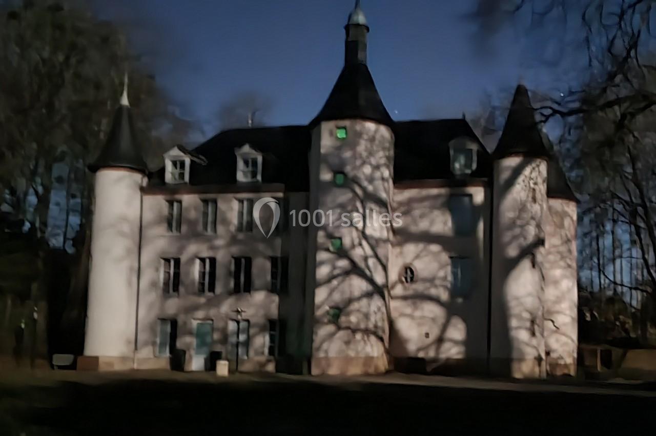 Château blanc entouré d'arbres, avec des ombres projetées sur la façade sous un ciel sombre.