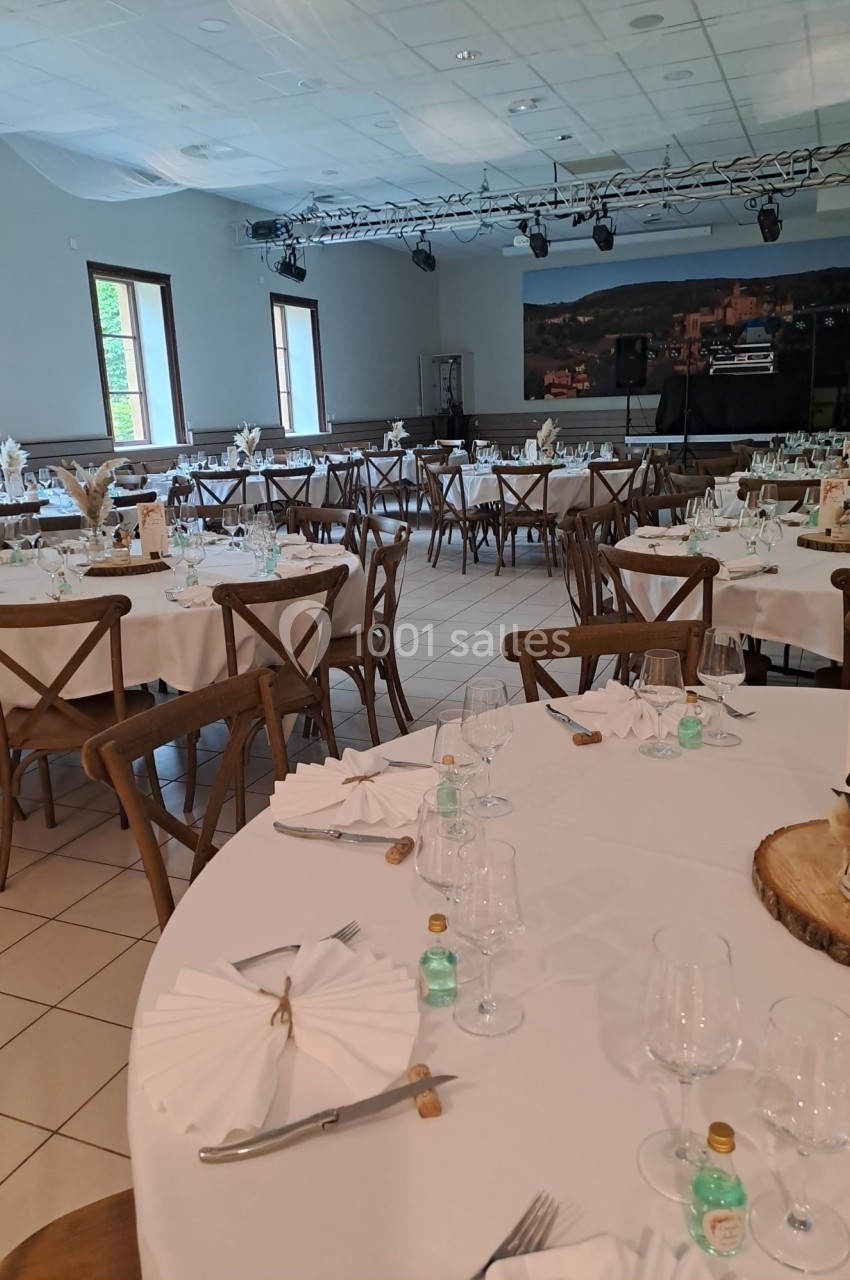 Salle de réception décorée avec des tables rondes dressées, chaises en bois et lumière naturelle entrant par les fenêtres.