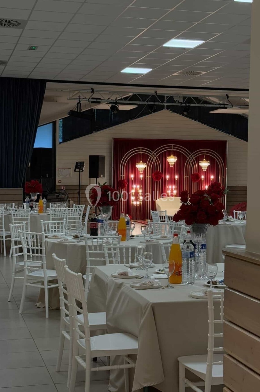 Salle de réception décorée avec des tables dressées, des chaises blanches et une scène ornée de lumières et de fleurs rouges.