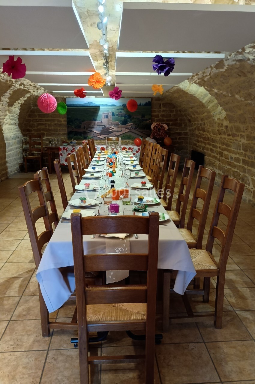 Table longue dressée pour un repas festif dans une salle voûtée en pierre, décorée de lanternes et guirlandes colorées.