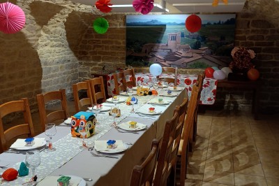 Salle de réception avec tables dressées, décorées de feuillages et vaisselle, éclairée par de grandes baies vitrées.
