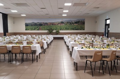 Salle de réception avec tables dressées, décorées de feuillages et vaisselle, éclairée par de grandes baies vitrées.