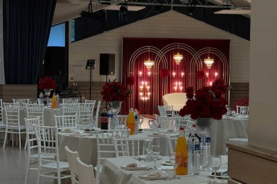 Salle de réception avec tables dressées, décorées de feuillages et vaisselle, éclairée par de grandes baies vitrées.