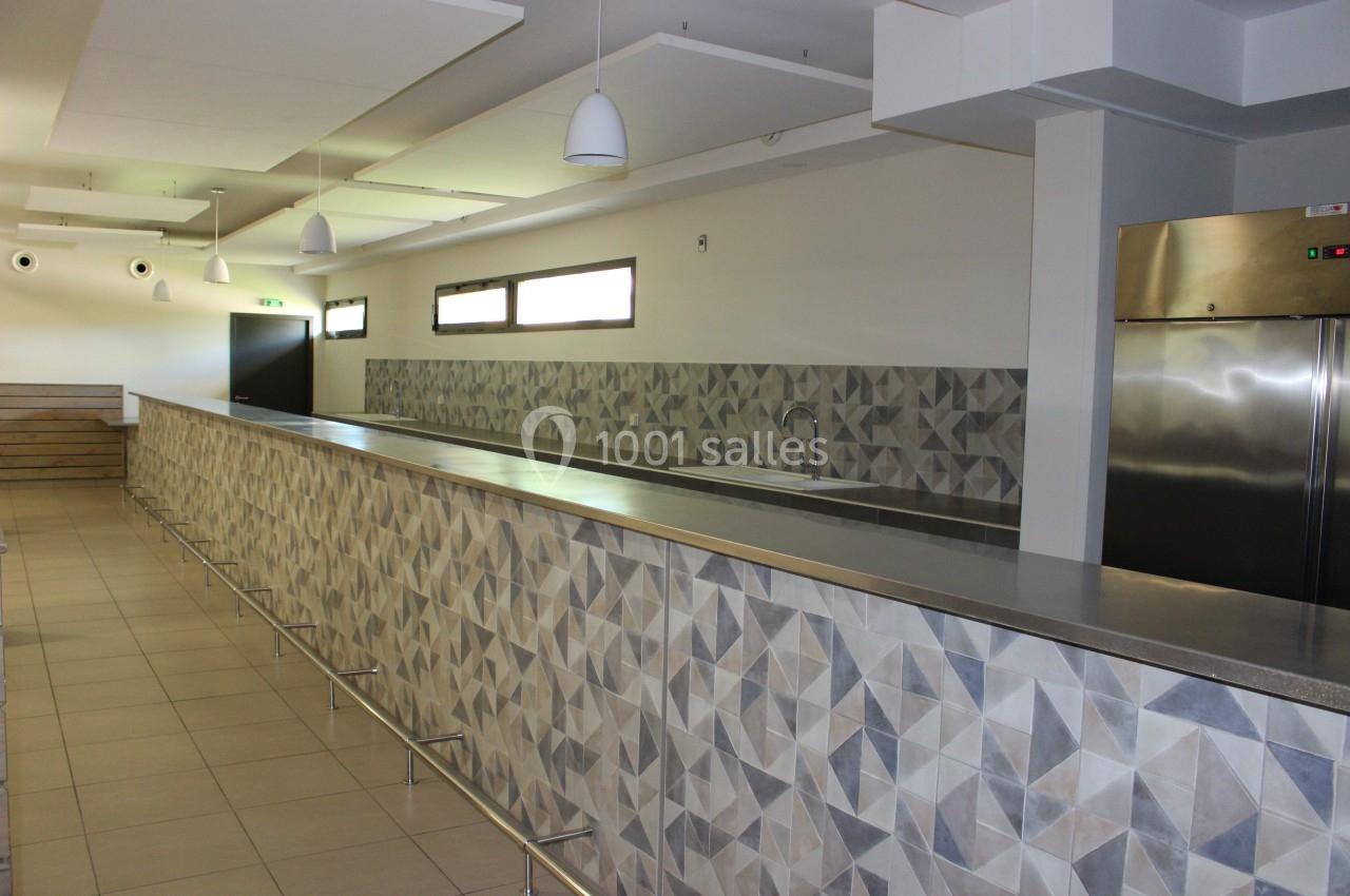 Comptoir long avec revêtement en carreaux géométriques, éclairage suspendu et réfrigérateur en acier inoxydable.