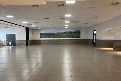 Salle lumineuse avec carrelage clair, baies vitrées, rangées de chaises empilées et un comptoir au fond.