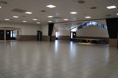 Salle lumineuse avec carrelage clair, baies vitrées, rangées de chaises empilées et un comptoir au fond.