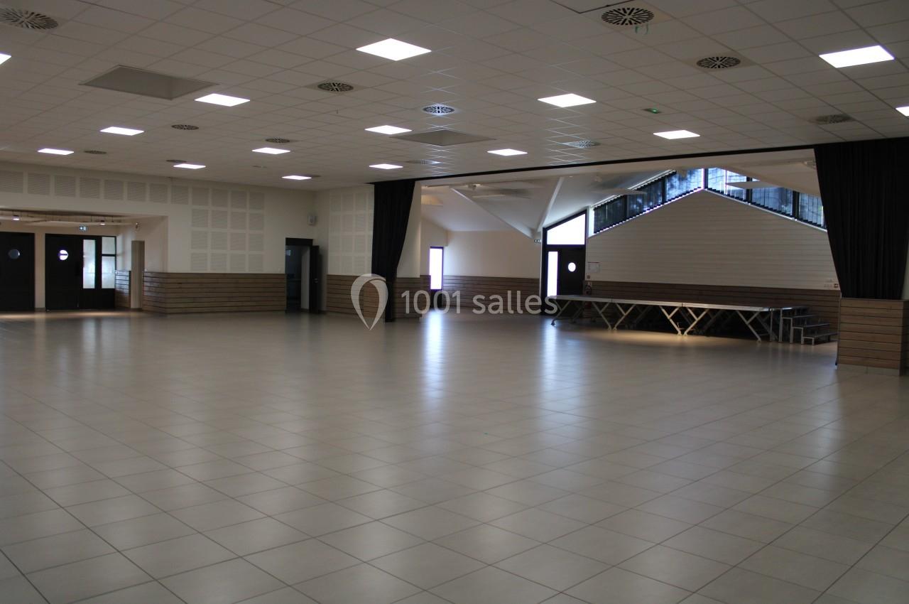 Salle spacieuse avec sol carrelé, murs clairs, plafonniers allumés et estrade en bois près d'une baie vitrée.