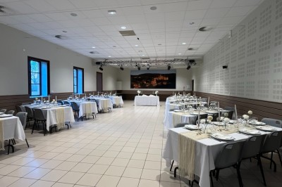 Salle de réception avec tables dressées, décorées de feuillages et vaisselle, éclairée par de grandes baies vitrées.