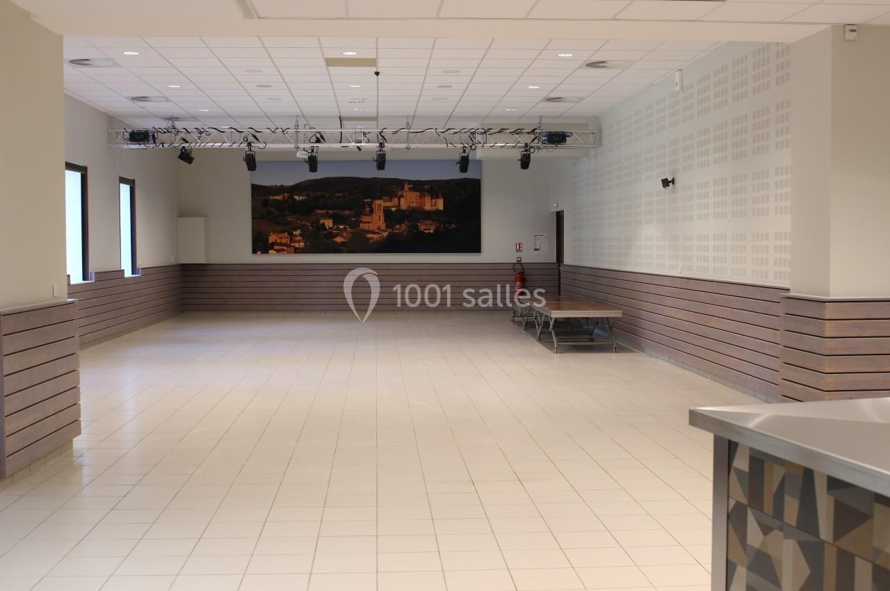 Salle polyvalente vide avec carrelage clair, murs blancs, éclairage suspendu et une scène en fond.