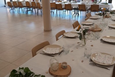 Salle de réception avec tables dressées, décorées de feuillages et vaisselle, éclairée par de grandes baies vitrées.