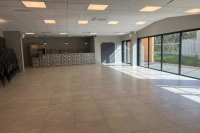 Salle lumineuse avec carrelage clair, baies vitrées, rangées de chaises empilées et un comptoir au fond.