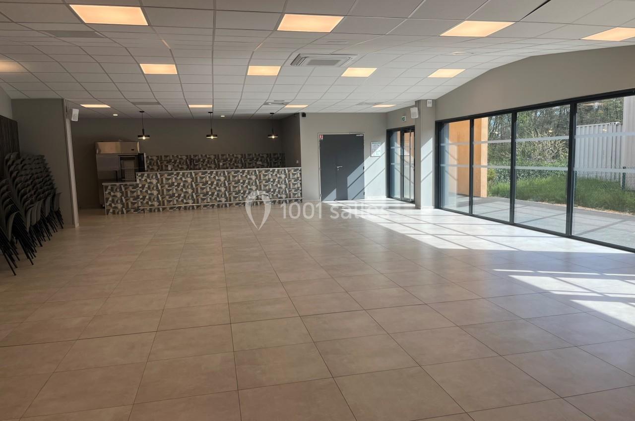 Salle lumineuse avec carrelage clair, baies vitrées, rangées de chaises empilées et un comptoir au fond.