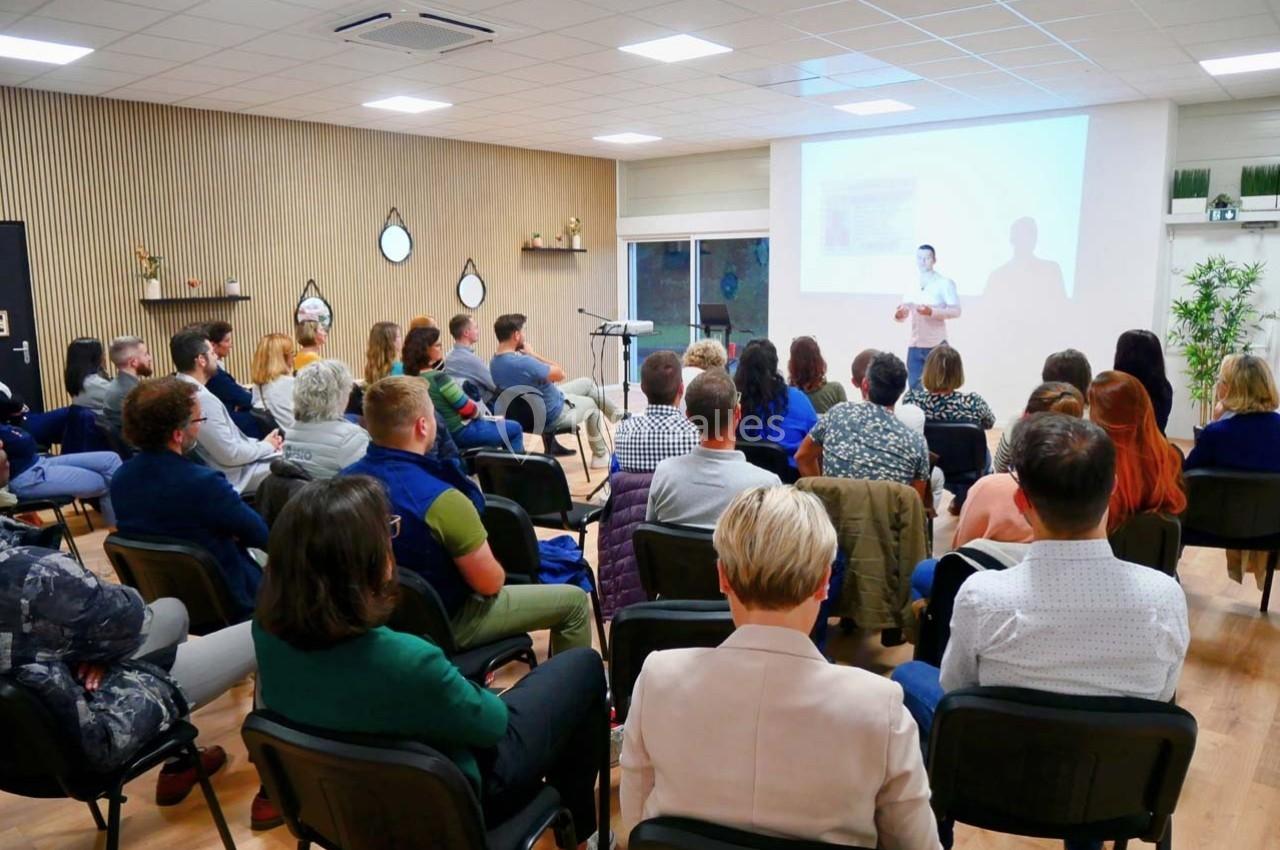Un conférencier s'adresse à un public assis dans une salle lumineuse avec un écran de projection.