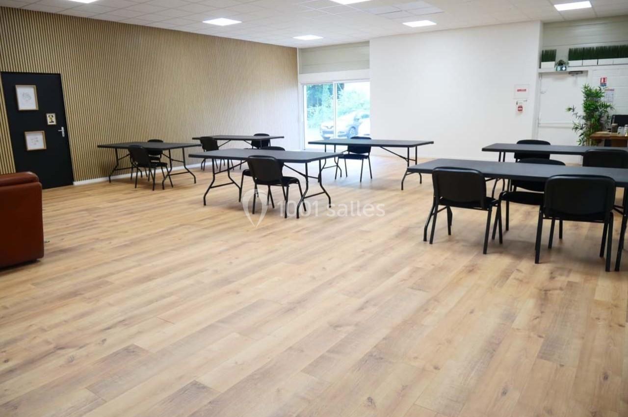 Salle lumineuse avec parquet, tables et chaises espacées, murs clairs et une baie vitrée donnant sur l'extérieur.