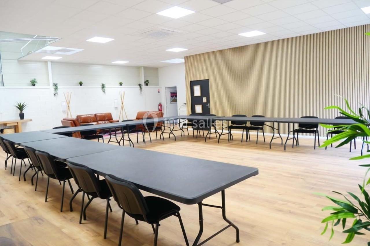 Salle lumineuse avec tables noires disposées en U, chaises noires, mur en bois clair et plantes décoratives.