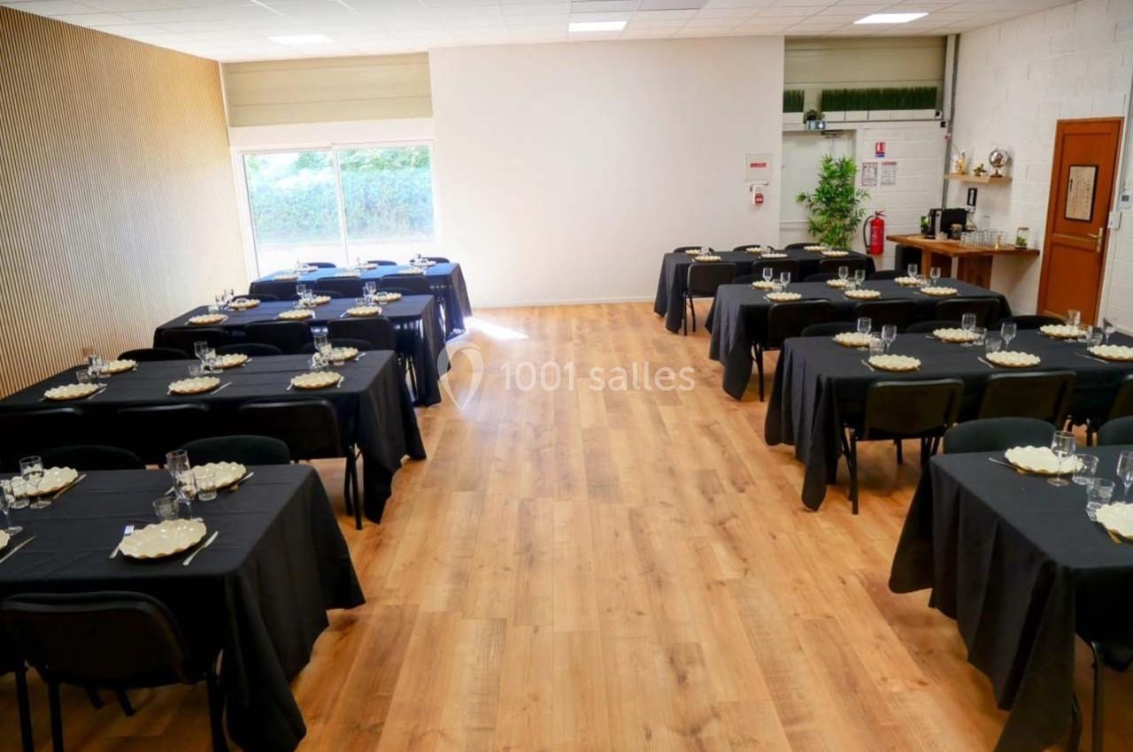 Salle lumineuse aménagée avec des tables noires dressées pour un repas, disposées en rangées sur un sol en bois.