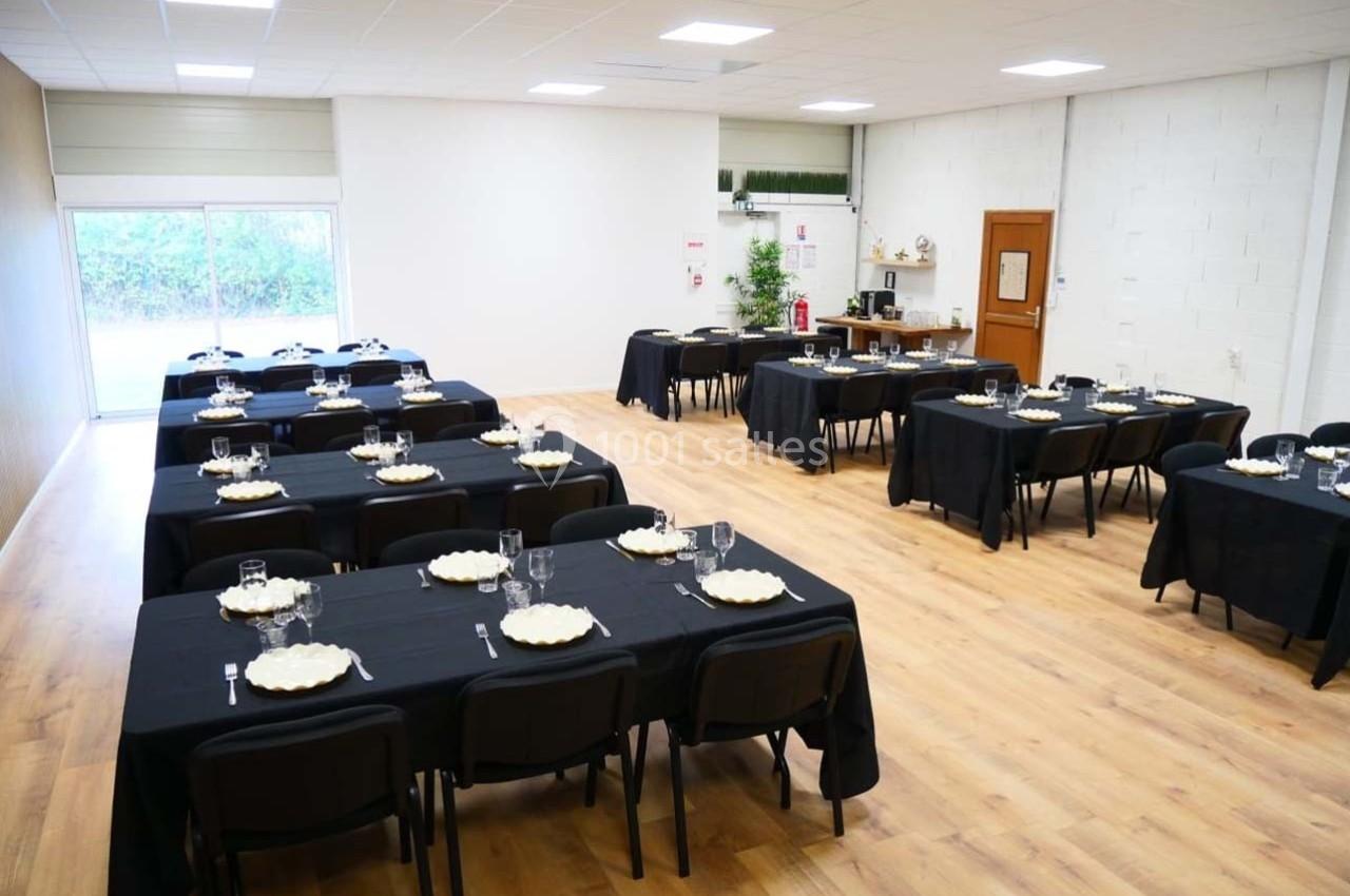 Salle de réception avec tables dressées en nappes noires, assiettes et verres, éclairée par une grande baie vitrée.