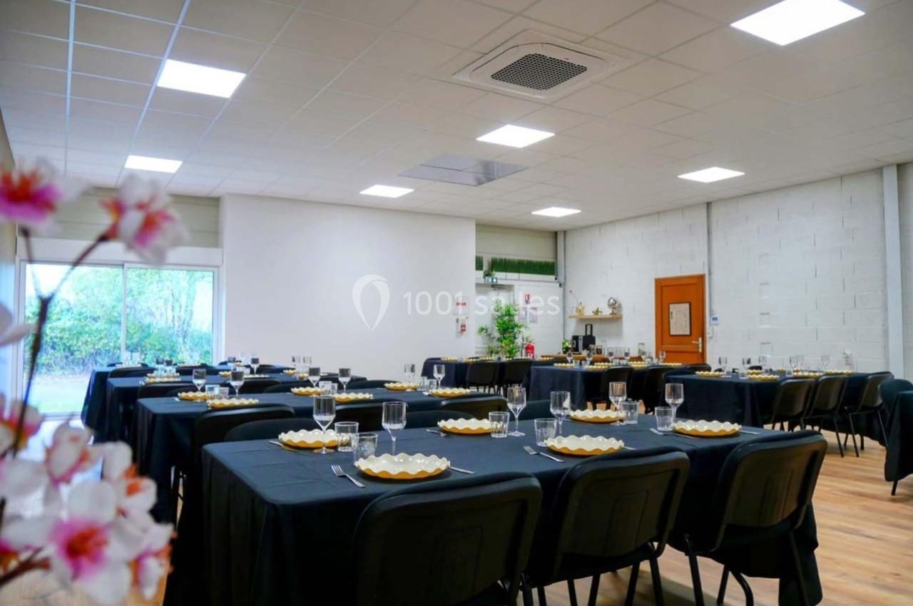 Salle lumineuse avec tables dressées de nappes noires, assiettes et verres, prête pour un repas ou un événement.