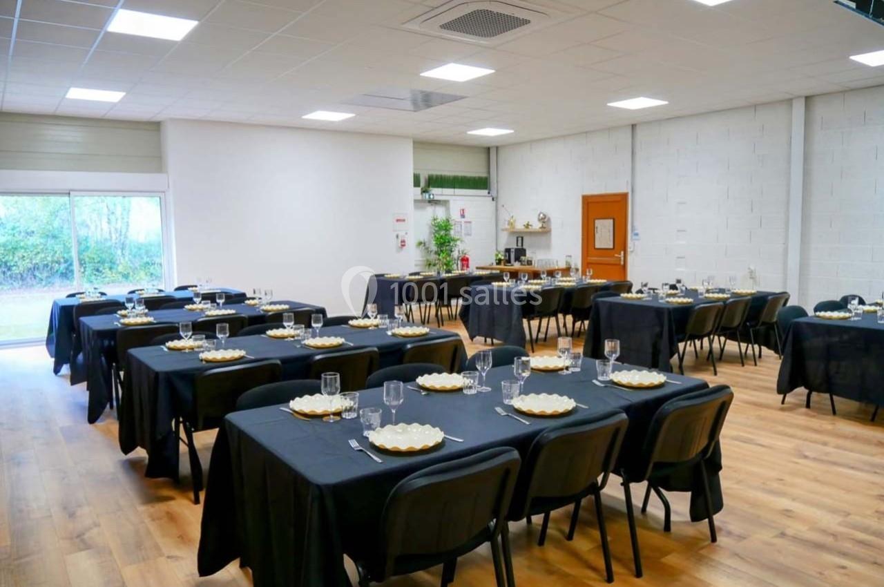 Salle aménagée avec des tables dressées de nappes noires, assiettes et verres, prête pour un événement ou un repas.