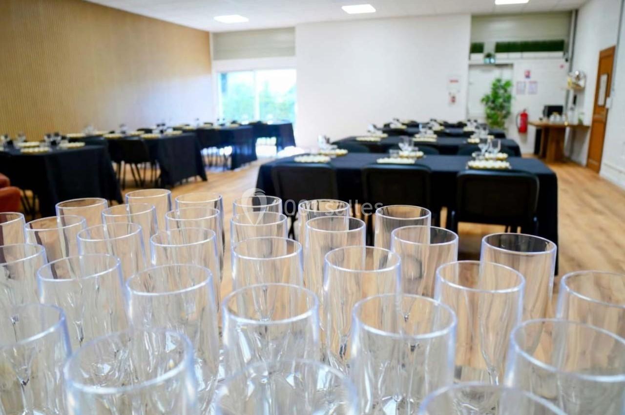 Salle de réception avec tables dressées en arrière-plan et rangée de verres vides au premier plan.