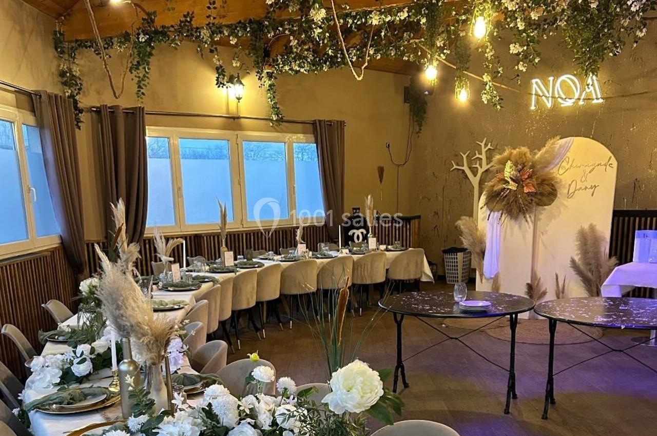 Salle décorée pour un événement avec tables dressées, fleurs blanches, guirlandes lumineuses et panneau lumineux ’NOA’.