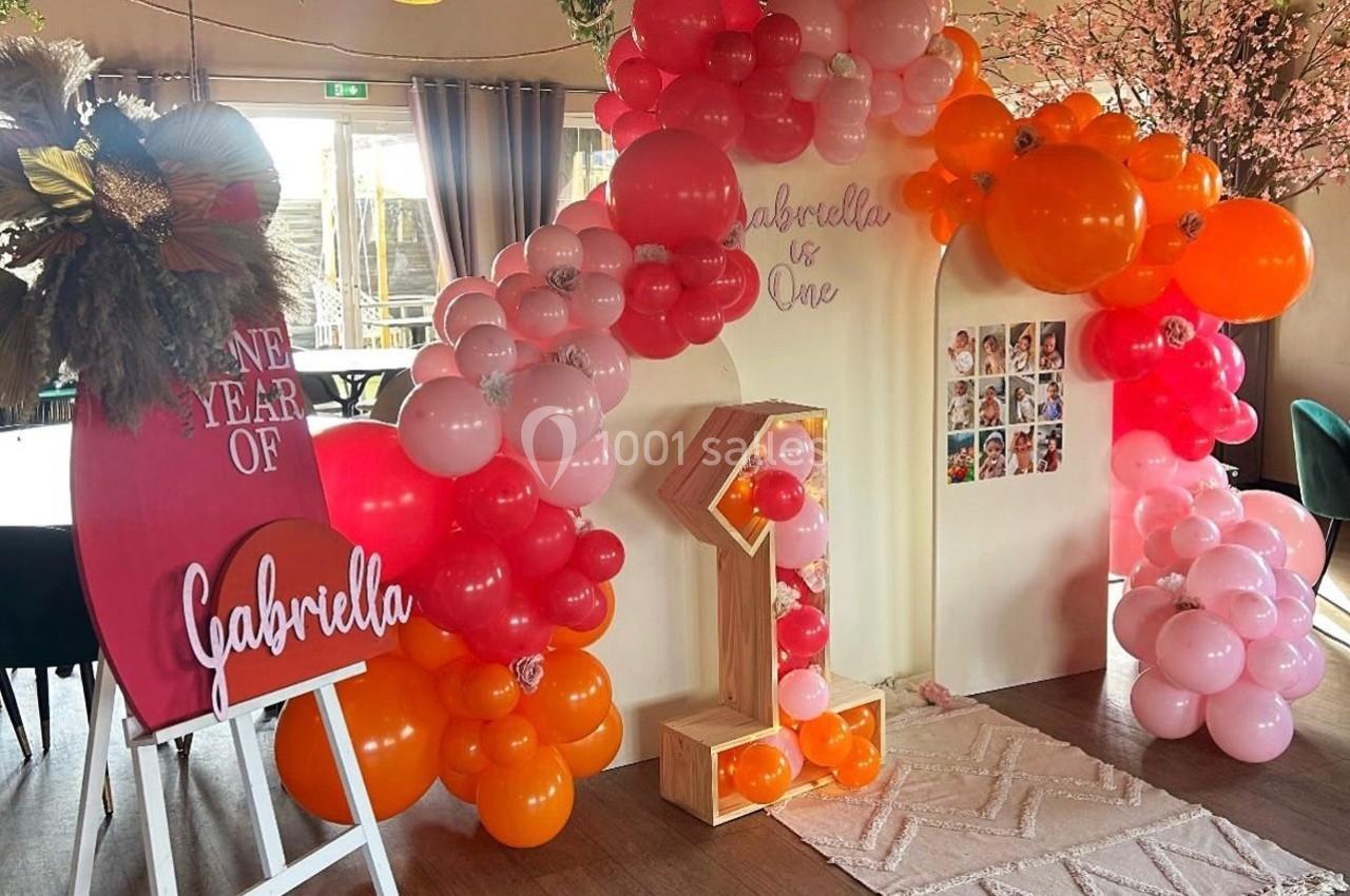 Décoration festive avec arche de ballons roses et oranges, chiffre ’1’ lumineux et panneau personnalisé pour un anniversaire.