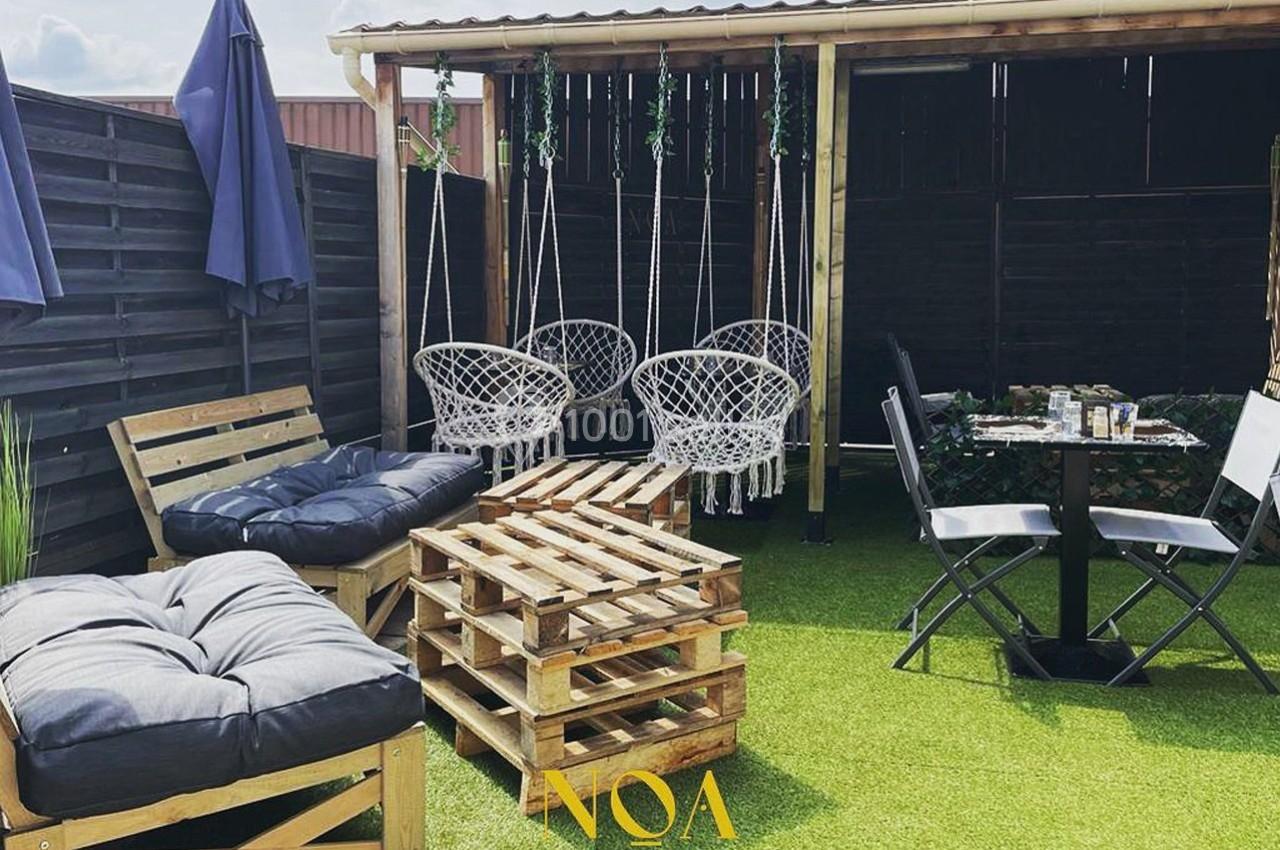 Espace extérieur aménagé avec fauteuils en palettes, balançoires suspendues, table et chaises sur gazon artificiel.