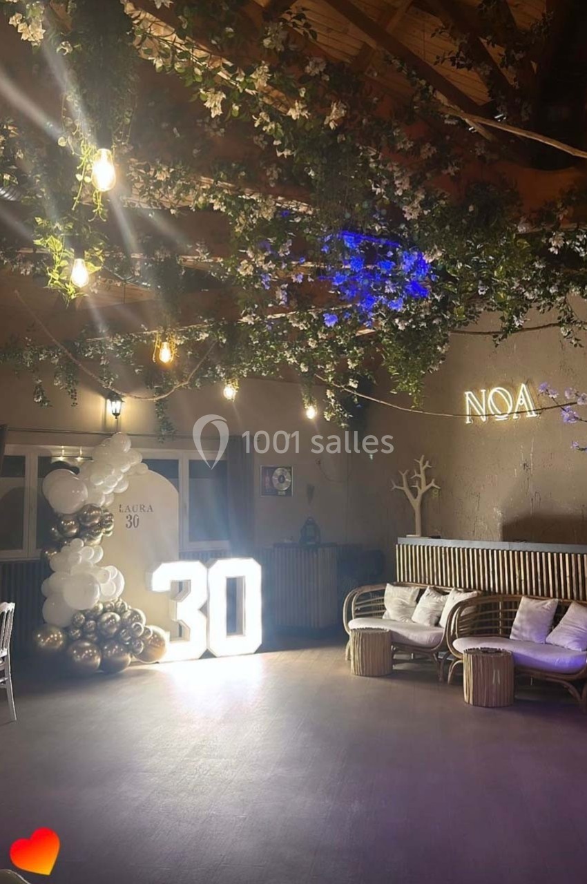 Décoration d'anniversaire avec ballons, chiffre lumineux ’30’, guirlandes végétales et néon ’Noa’ dans une salle.