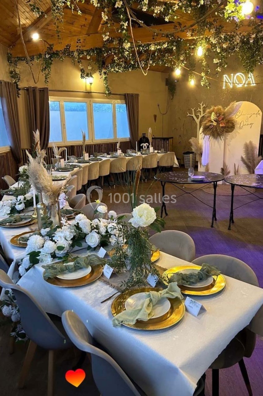 Salle décorée pour un événement avec tables dressées, assiettes dorées, fleurs blanches et éclairage chaleureux.